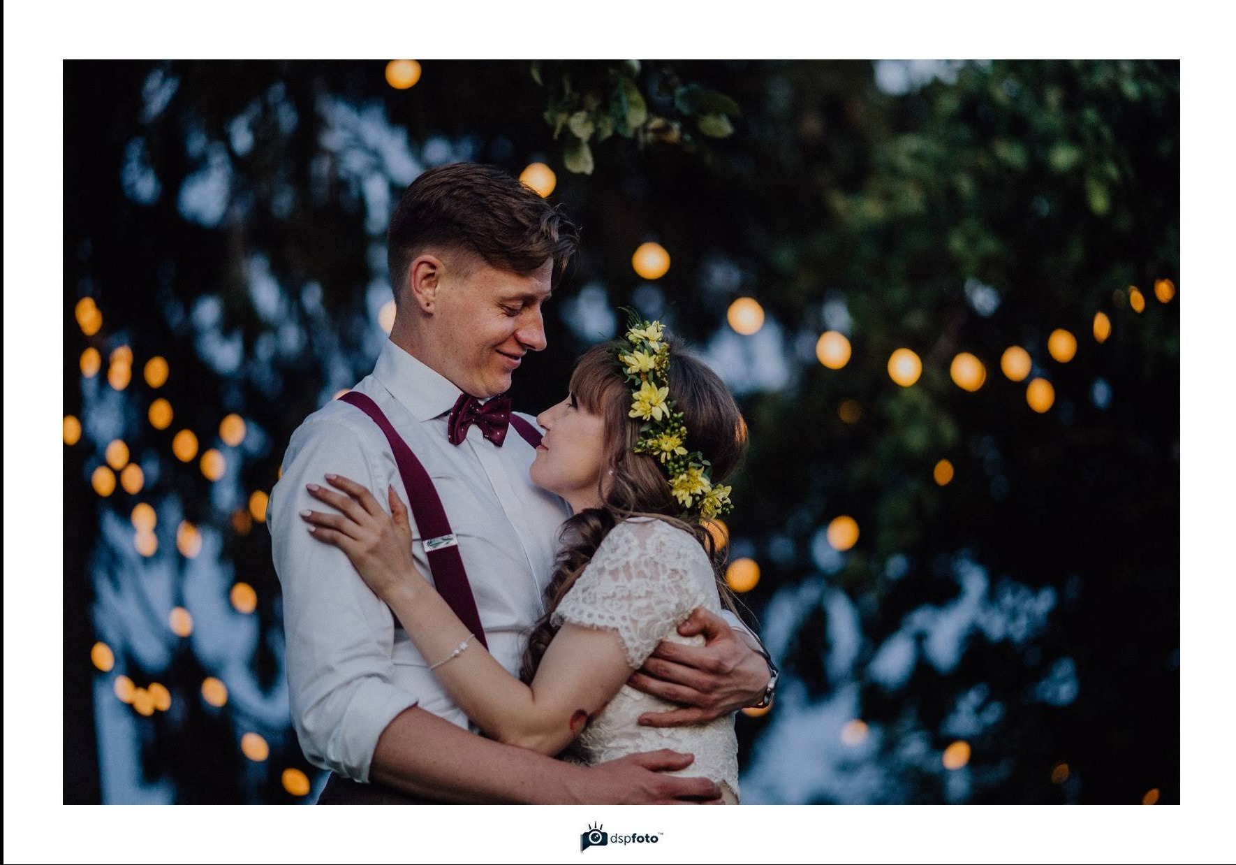 Uśmiechnięta para młoda w objęciach, panna młoda z wiankiem z żółtych kwiatów, pan młody w białej koszuli, bordowych szelkach i muszce, w tle rozproszone ciepłe światła