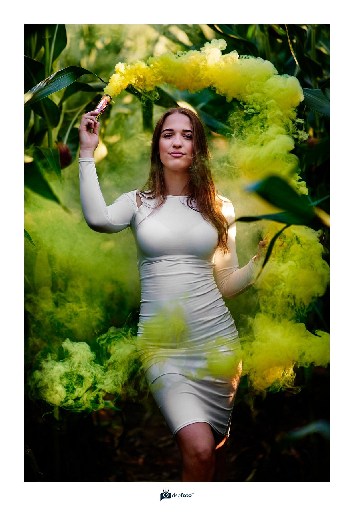 Młoda kobieta w białej sukience pozuje z żółtą świecą dymną w polu kukurydzy, otoczona gęstym dymem, z delikatnym uśmiechem na twarzy.