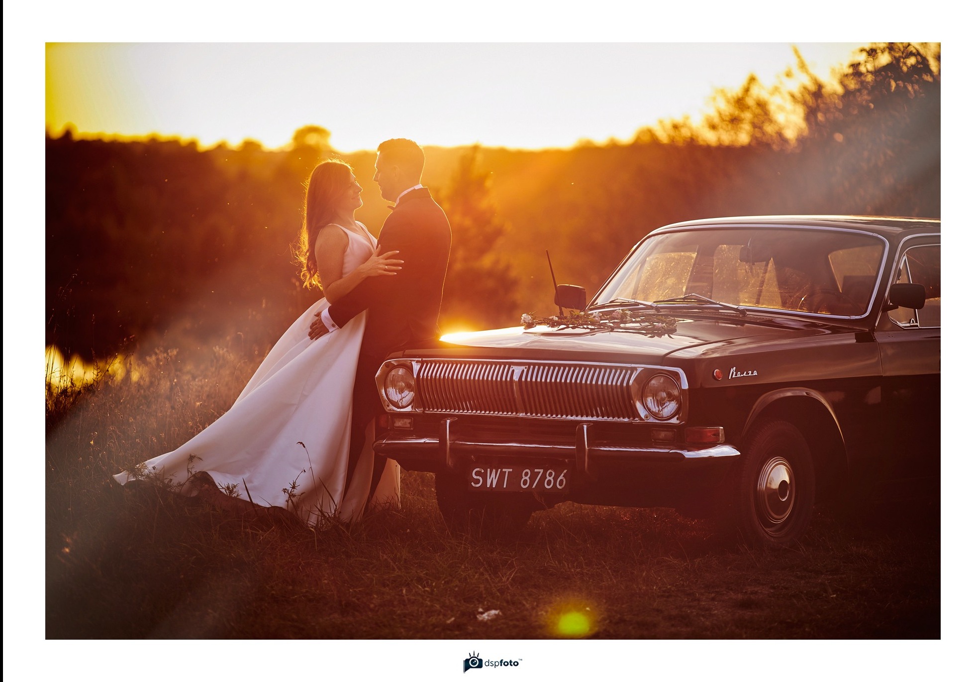 Para młoda w objęciach przy ciemnym samochodzie marki Wołga z kwiatami na masce, w tle zachód słońca