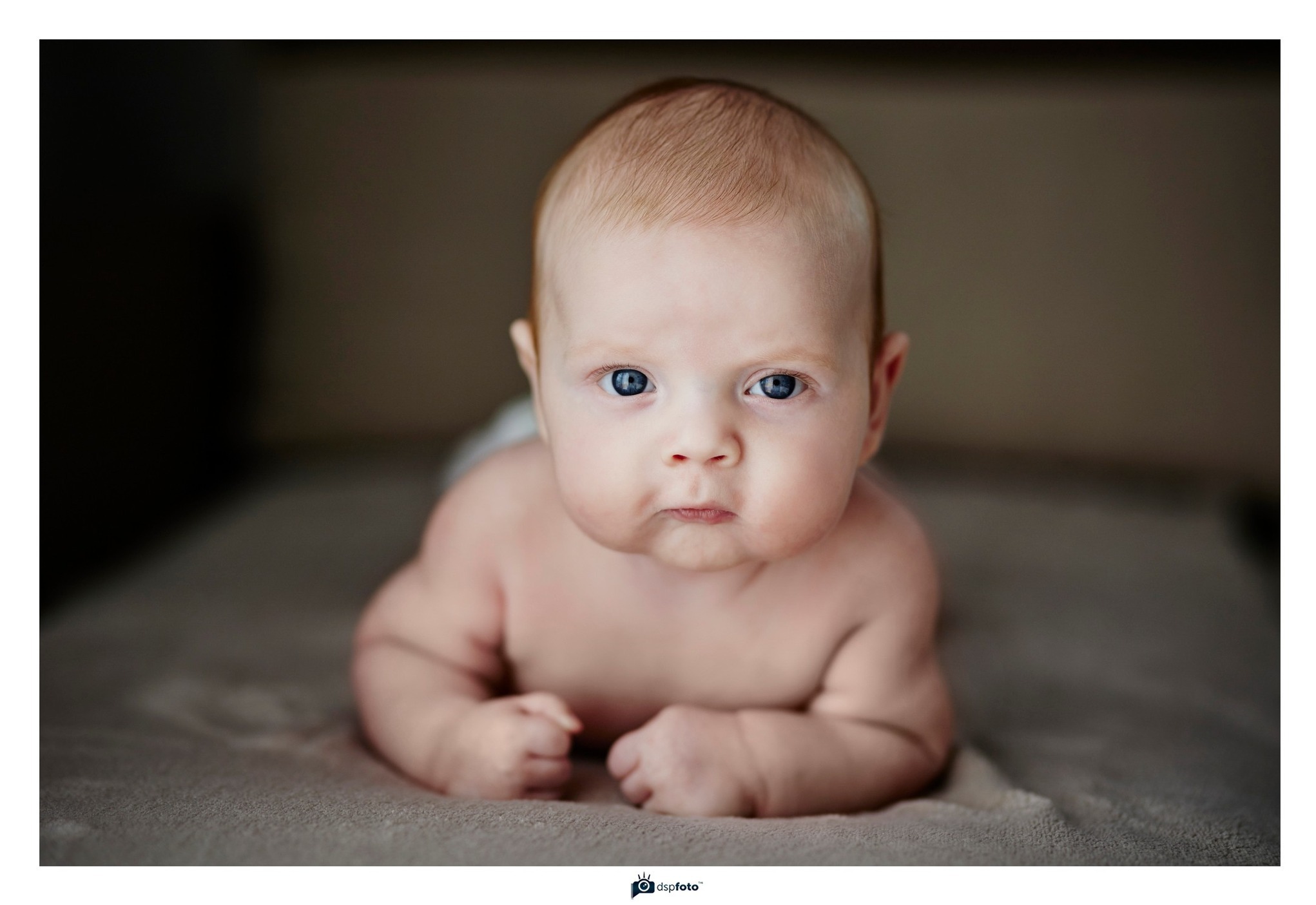 Portret noworodka leżącego na brzuchu, z intensywnym spojrzeniem niebieskich oczu i drobnymi, zaciśniętymi piąstkami.