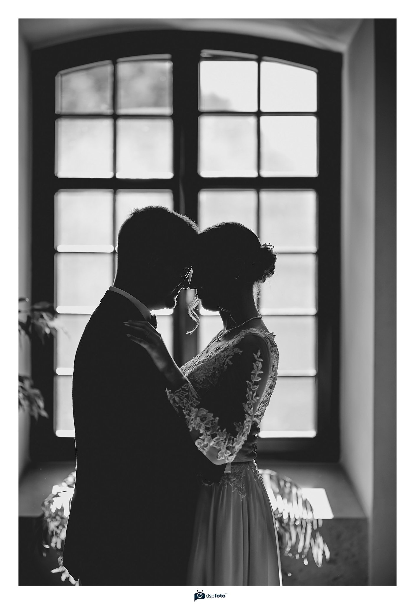 Czarno-biała fotografia pary młodej w objęciach na tle okna z kratami, delikatne światło podkreśla koronkowy detal sukni panny młodej.