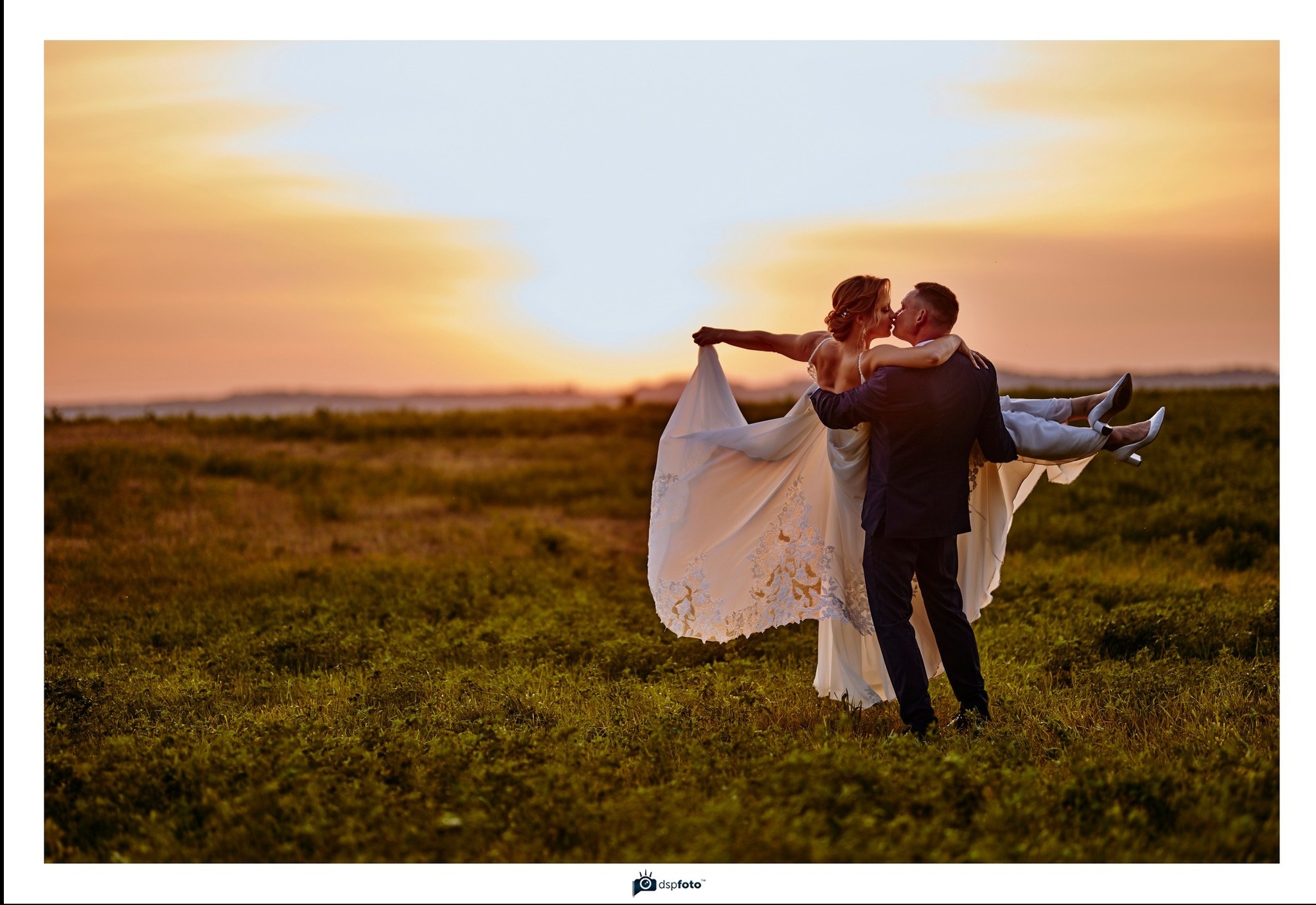 Pan młody trzyma pannę młodą na rękach na tle zachodzącego słońca, panna młoda w białej sukni z koronkowymi zdobieniami i białych szpilkach.