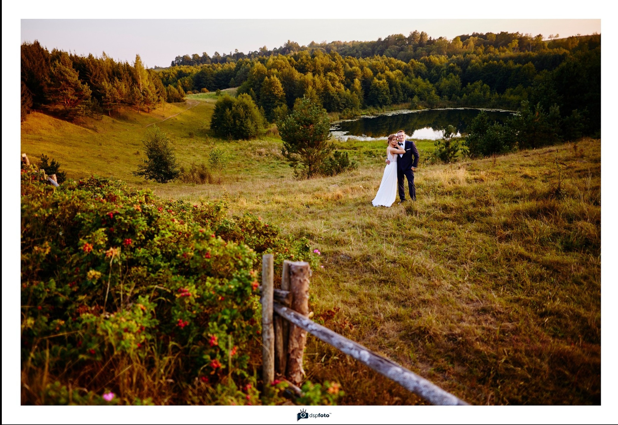 Para młoda w plenerze na wzgórzu z jeziorem w tle, ujęcie zza drewnianego płotu i kwitnących krzewów.