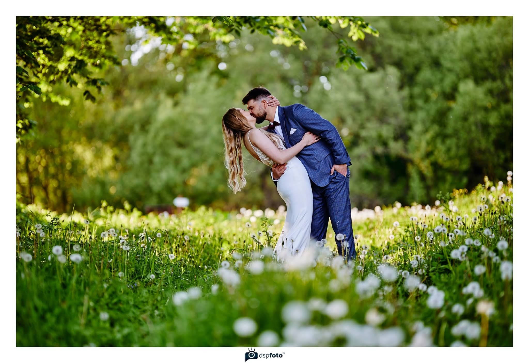 Para młoda w plenerze: on w granatowym garniturze w kratę, ona w białej sukni, całują się na łące z dmuchawcami, w tle zieleń drzew. Romantyczny kadr.