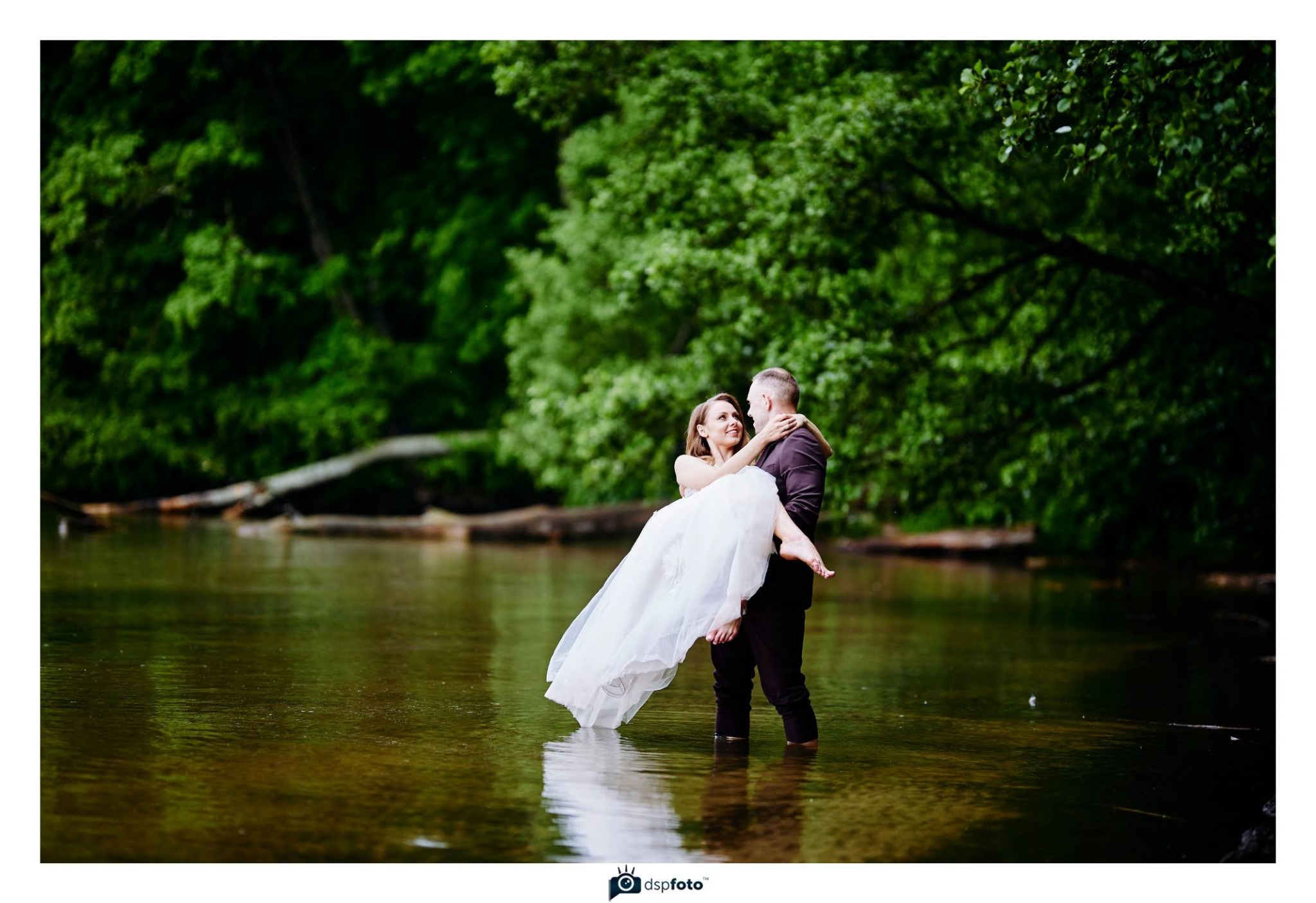 Uśmiechnięta panna młoda w białej sukni niesiona przez pana młodego w wodzie na tle zieleni drzew. Romantyczna sesja plenerowa w naturalnym otoczeniu.
