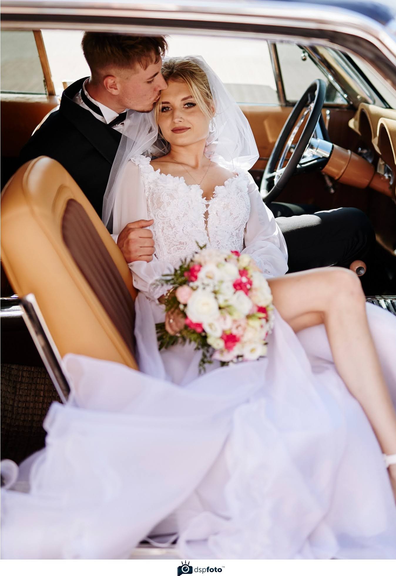 Para młoda w klasycznym samochodzie: panna młoda w białej sukni z bukietem, pan młody w smokingu całuje ją w czoło. Skórzana tapicerka i kierownica auta.