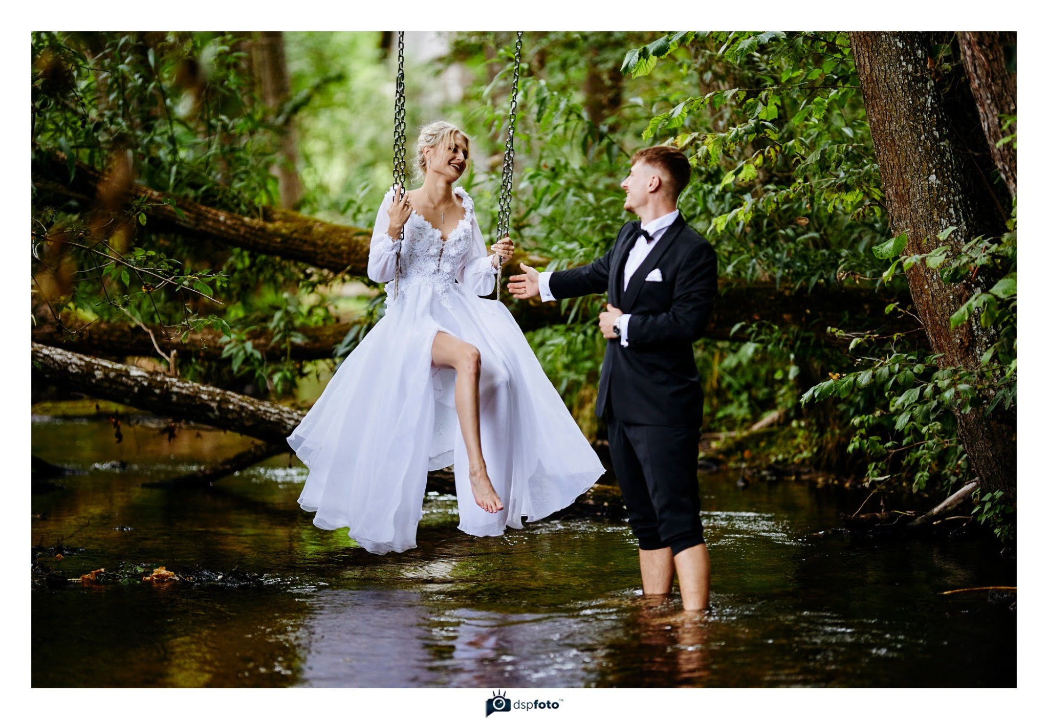 Uśmiechnięta panna młoda na huśtawce nad rzeką, w tle las, obok pan młody w wodzie. Nietypowa sesja ślubna w plenerze, naturalne światło.