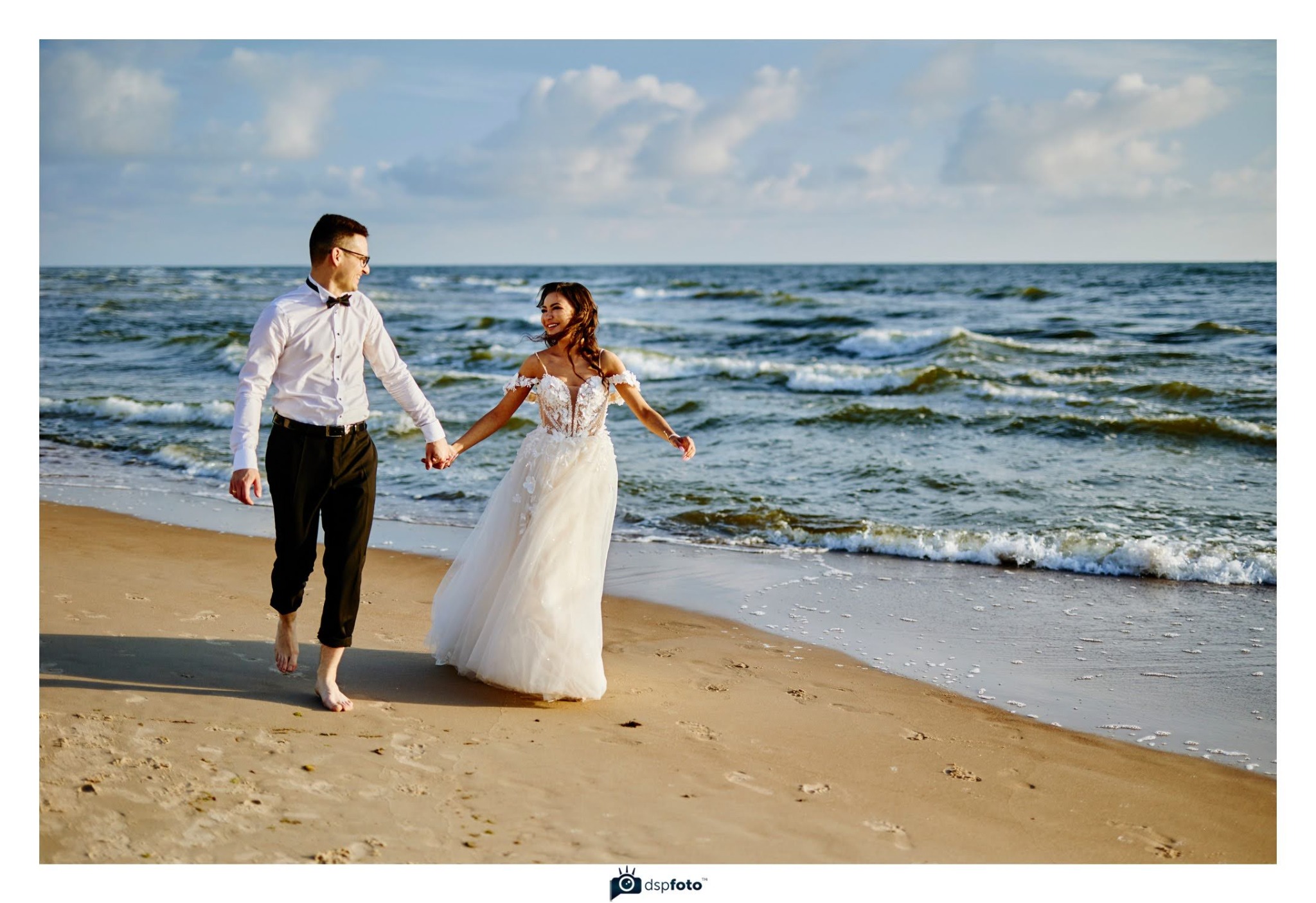 Para młoda, boso, idzie brzegiem morza trzymając się za ręce. Panna młoda w białej sukni, pan młody w białej koszuli i czarnych spodniach. Słońce, fale, piasek.
