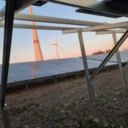 Panel słoneczny na konstrukcji stalowej, w tle turbiny wiatrowe oświetlone zachodzącym słońcem.