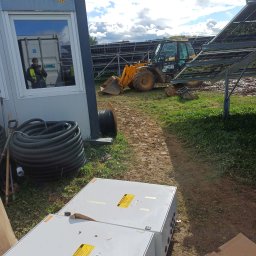 Dziurzyński Kacper G4 free Energy - Plac budowy z traktorem JCB, rozłożonymi konstrukcjami pod panele i białymi skrzynkami elektrycznymi; odbicie pracownika w oknie kontenera budowlanego. Widoczne zwoje kabli i narzędzia.