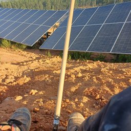 Dziurzyński Kacper G4 free Energy - Relaks podczas montażu: Widok z perspektywy osoby siedzącej na ziemi, z nogami w roboczych butach i narzędziem wbitą w czerwoną glebę, w tle panele słoneczne i las.