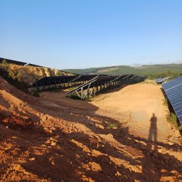 Dziurzyński Kacper G4 free Energy - Montaż konstrukcji wsporczej dla paneli fotowoltaicznych na nierównym, piaszczystym terenie z widocznym cieniem osoby w słoneczny dzień.