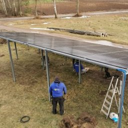 Dziurzyński Kacper G4 free Energy - Montaż paneli słonecznych na metalowej konstrukcji w terenie niezabudowanym, widoczni dwaj instalatorzy w niebieskich bluzach z logo firmy, drabina oparta o konstrukcję, wiosenna sceneria.