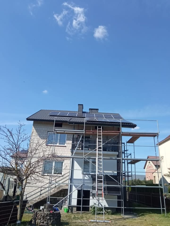 Dom dwupiętrowy w trakcie montażu paneli słonecznych na dachu, widoczne rusztowanie i drabina oparta o ścianę budynku.