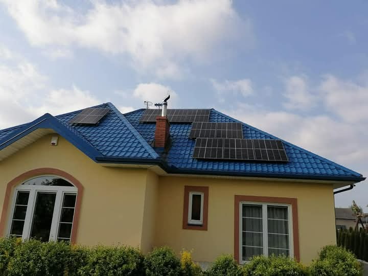 Dom jednorodzinny z niebieską blachodachówką i panelami słonecznymi na dachu, widok z poziomu ogrodu, jasne niebo z chmurami w tle.