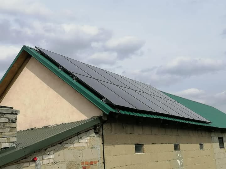 Panele słoneczne na zielonym dachu budynku gospodarczego z ceglanym kominem w pochmurny dzień.