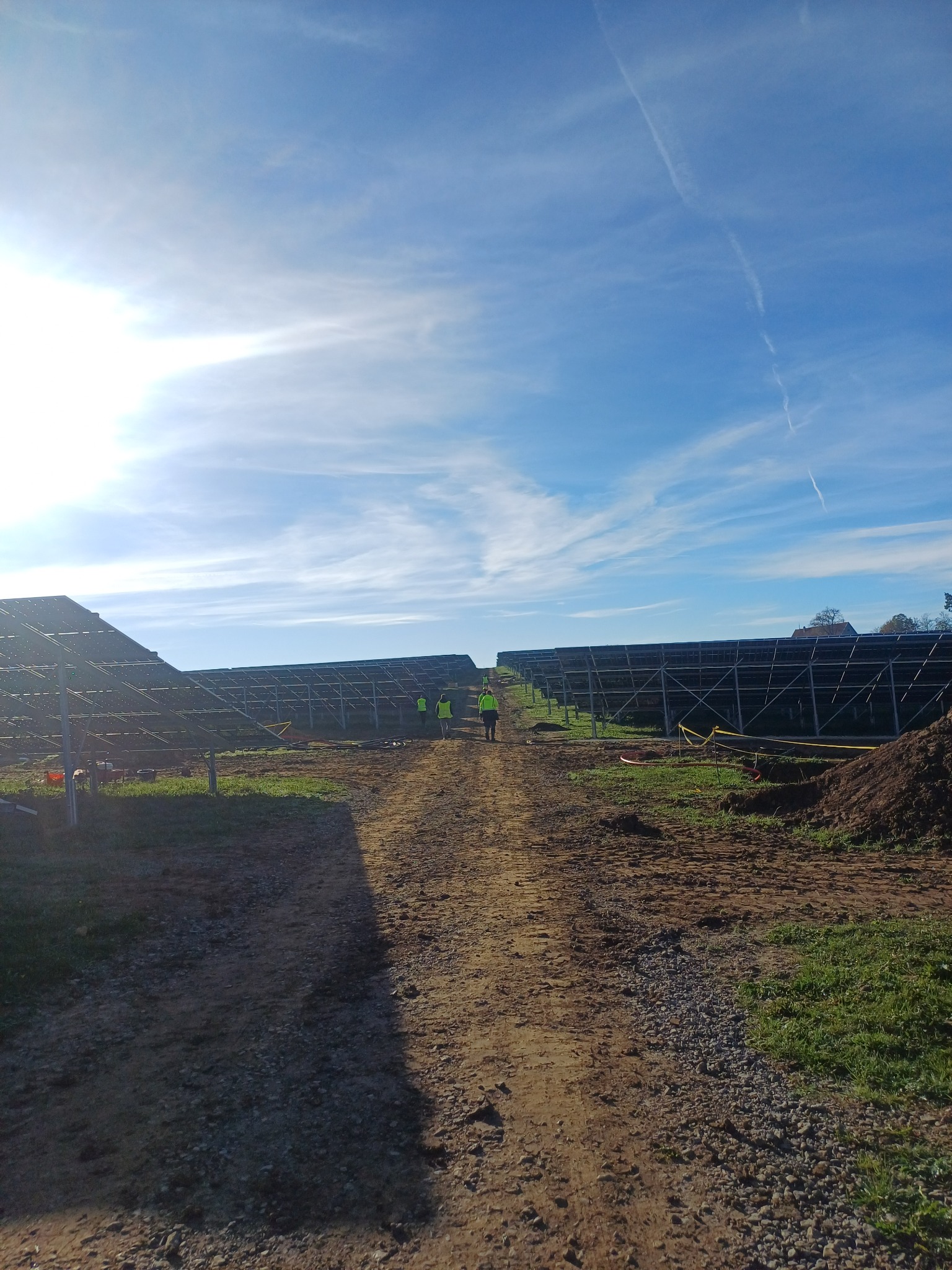 Rozległa instalacja paneli słonecznych w trakcie montażu, widoczna z perspektywy gruntu, z dwoma pracownikami w odblaskowych kamizelkach oddalającymi się ścieżką w kierunku środka kadru, błękitne...