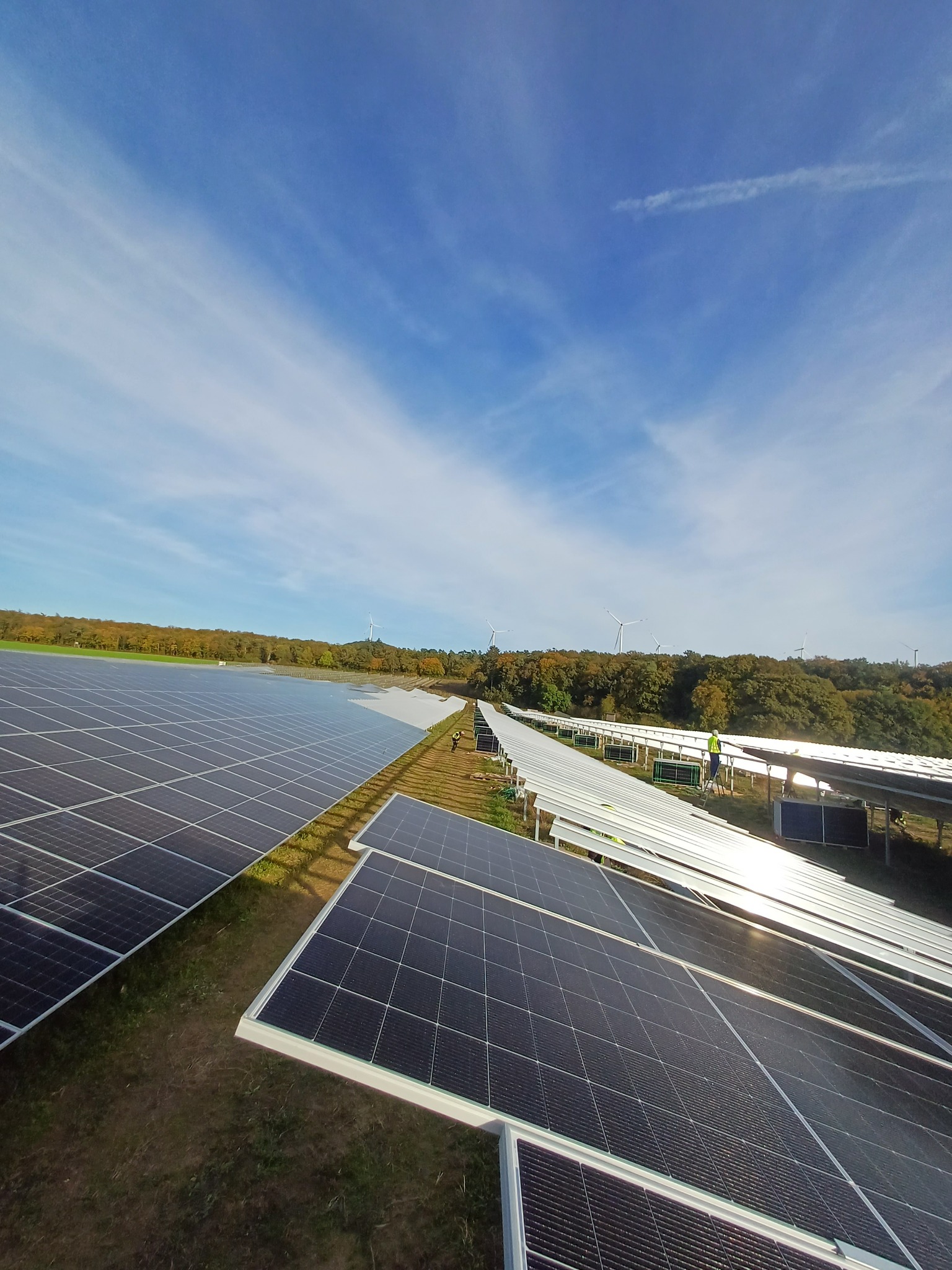 Rozległa farma paneli słonecznych w słoneczny dzień z widocznymi turbinami wiatrowymi w tle, pracownik na drabinie przy panelach.