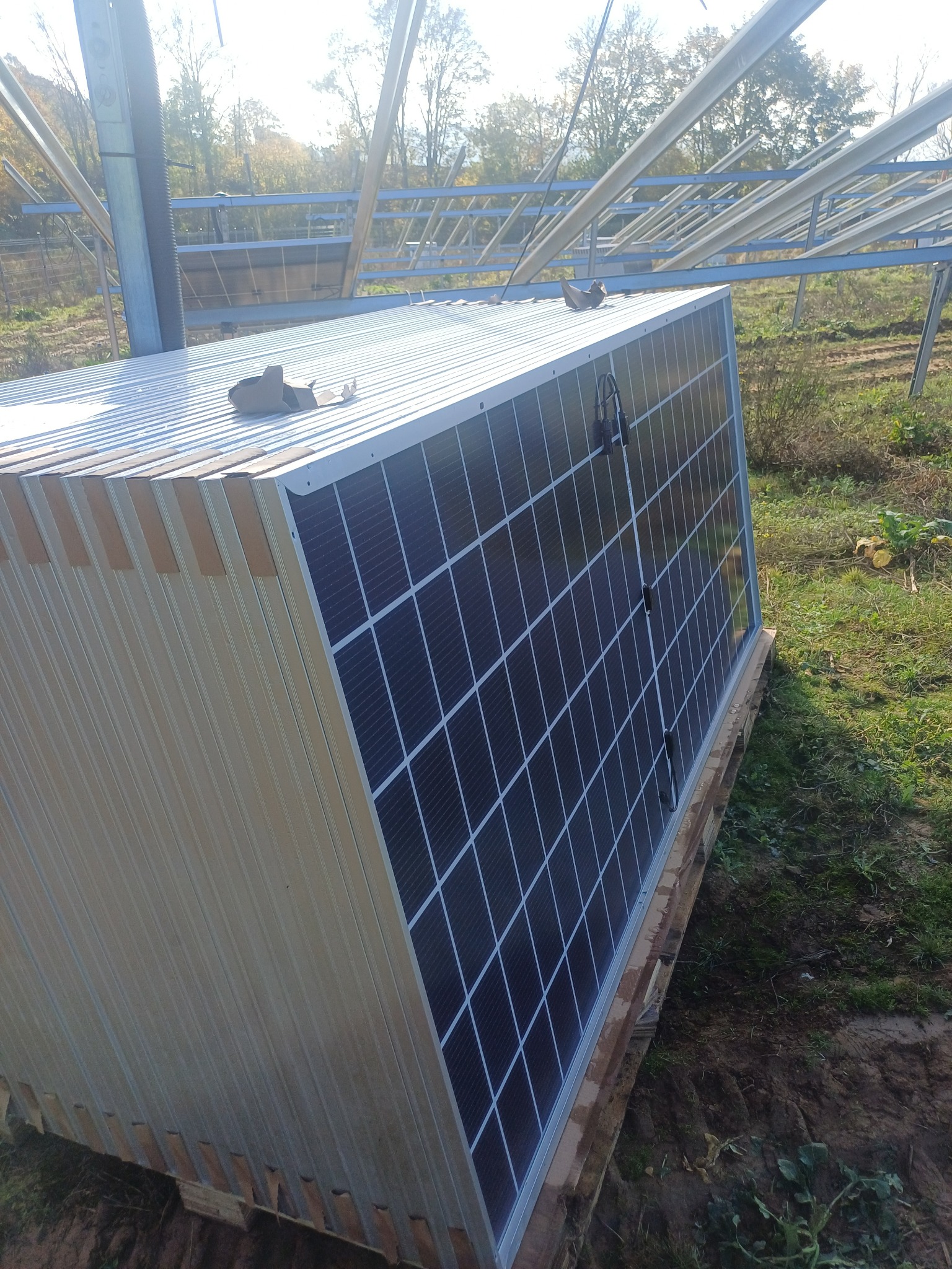 Nietypowa instalacja fotowoltaiczna: moduły zamontowane na blaszanym, prostopadłościennym obiekcie, prawdopodobnie magazynie energii, z widoczną konstrukcją wsporczą w tle i elementami krajobrazu.