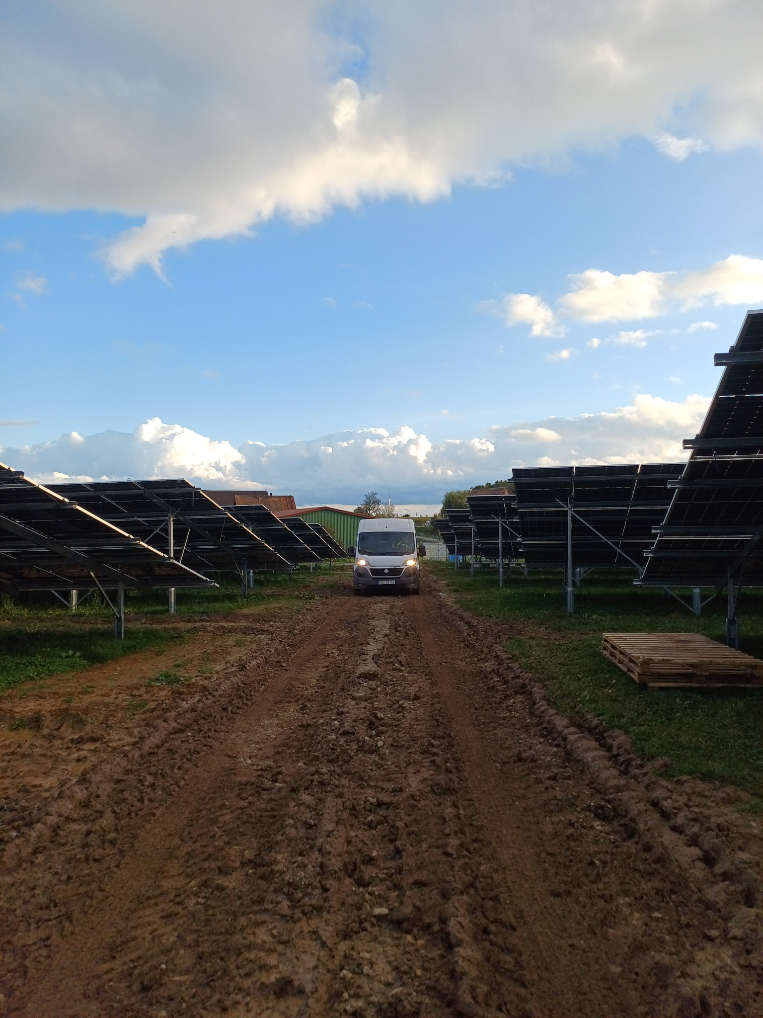 Dostawczy van na błotnistej drodze dojazdowej do rzędów paneli słonecznych na łące, pod błękitnym niebem z białymi chmurami.