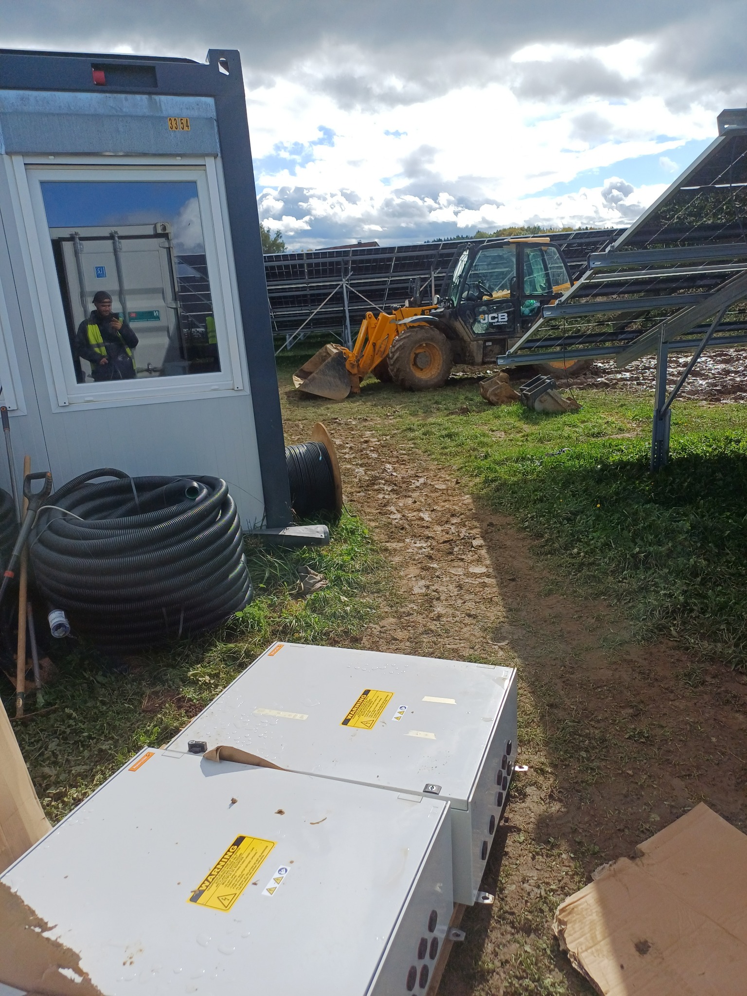 Plac budowy z traktorem JCB, rozłożonymi konstrukcjami pod panele i białymi skrzynkami elektrycznymi; odbicie pracownika w oknie kontenera budowlanego. Widoczne zwoje kabli i narzędzia.