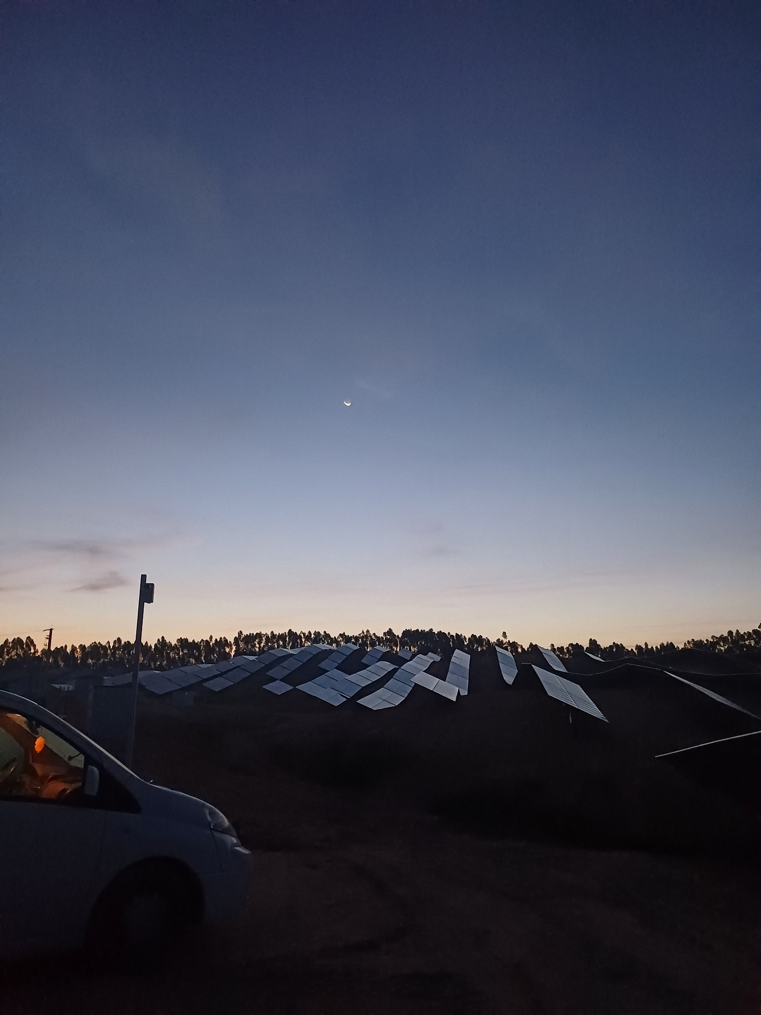 Instalacja paneli słonecznych na wzgórzu o zmierzchu, widoczny samochód serwisowy z pomarańczowym światłem w kabinie i cienki sierp księżyca na niebie.