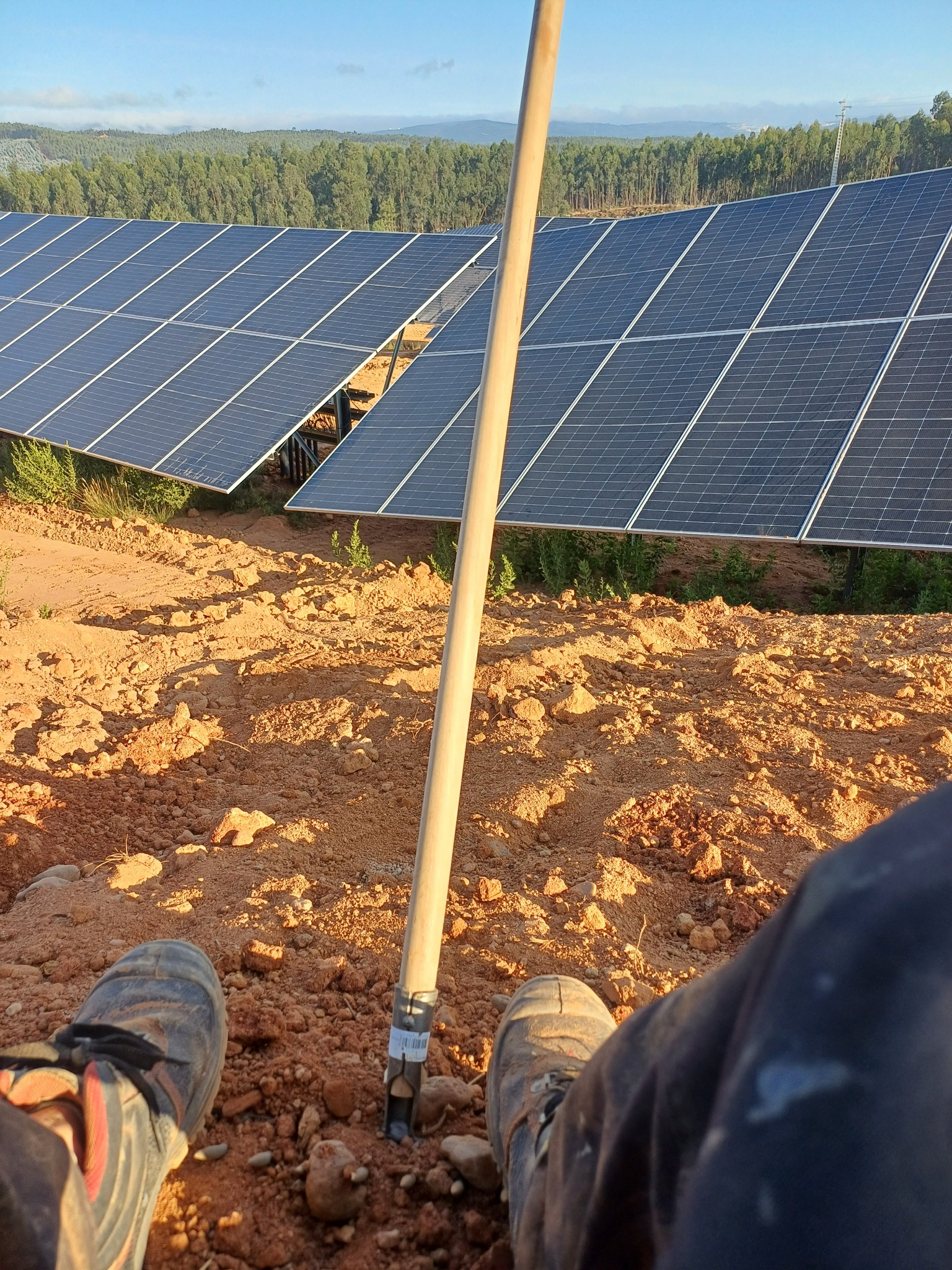 Relaks podczas montażu: Widok z perspektywy osoby siedzącej na ziemi, z nogami w roboczych butach i narzędziem wbitą w czerwoną glebę, w tle panele słoneczne i las.