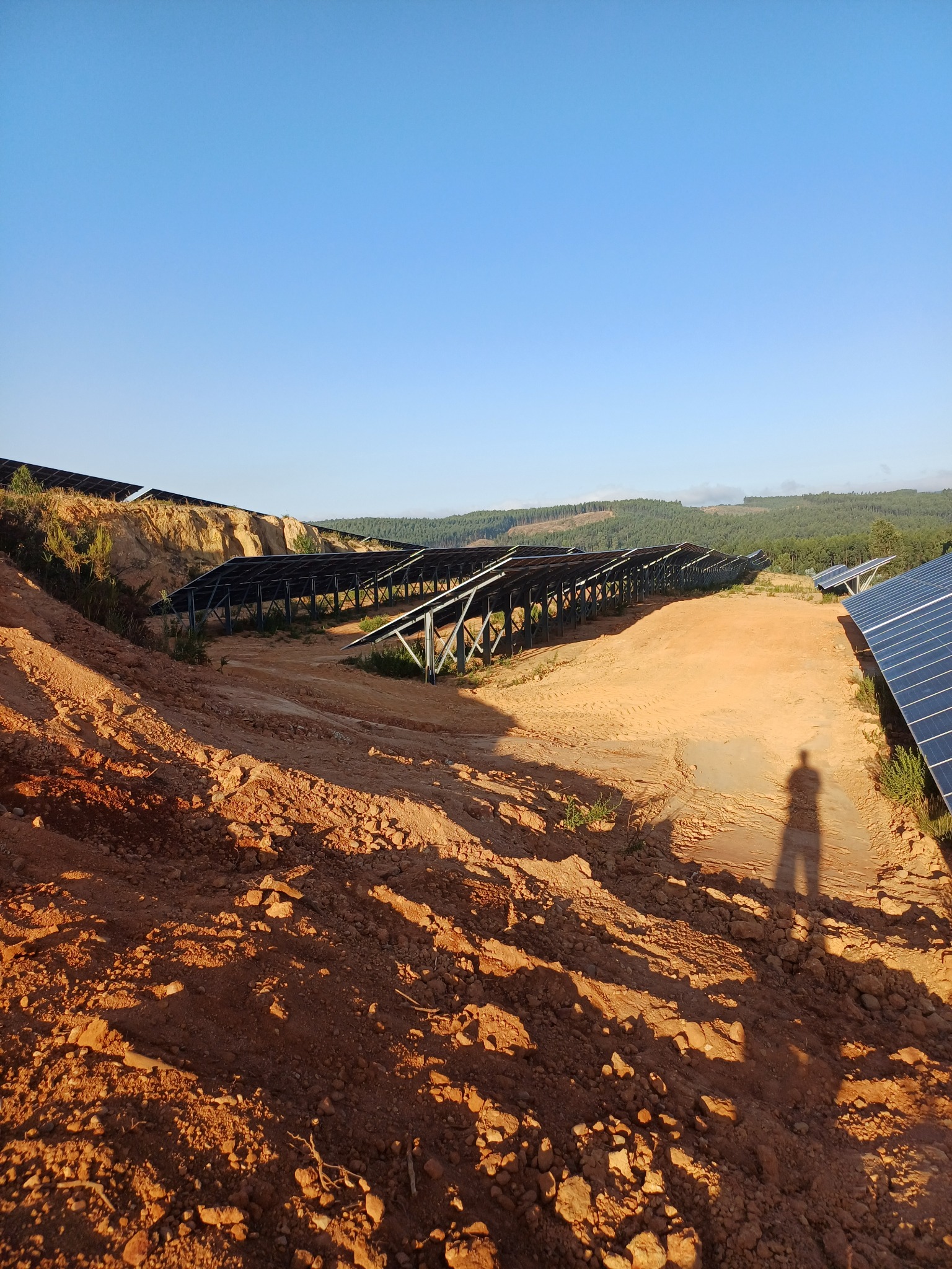 Montaż konstrukcji wsporczej dla paneli fotowoltaicznych na nierównym, piaszczystym terenie z widocznym cieniem osoby w słoneczny dzień.