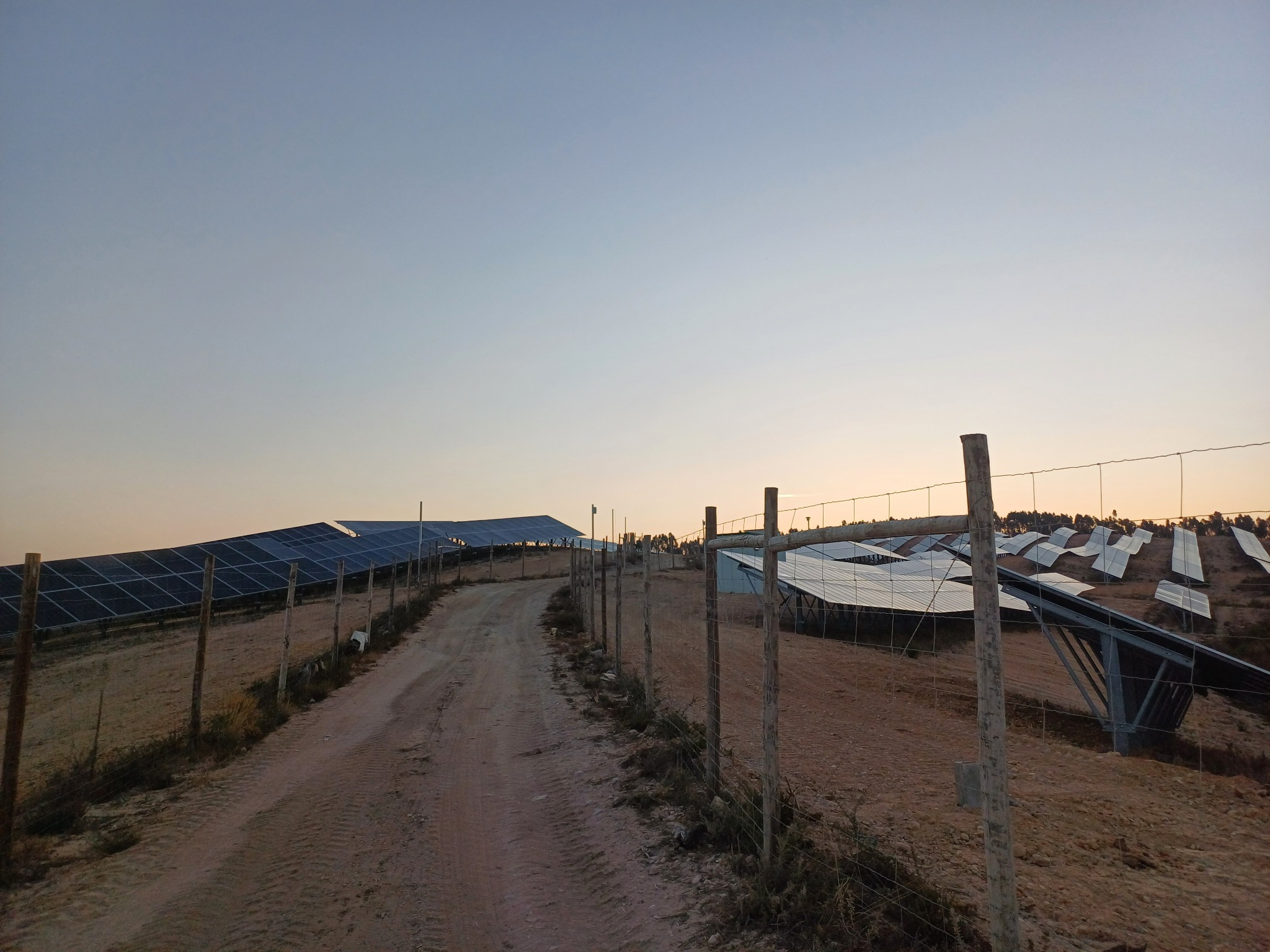 Rozległa farma paneli słonecznych na wzgórzu, widoczna z polnej drogi ogrodzonej drewnianym płotem z siatki, w tle zachodzące słońce.