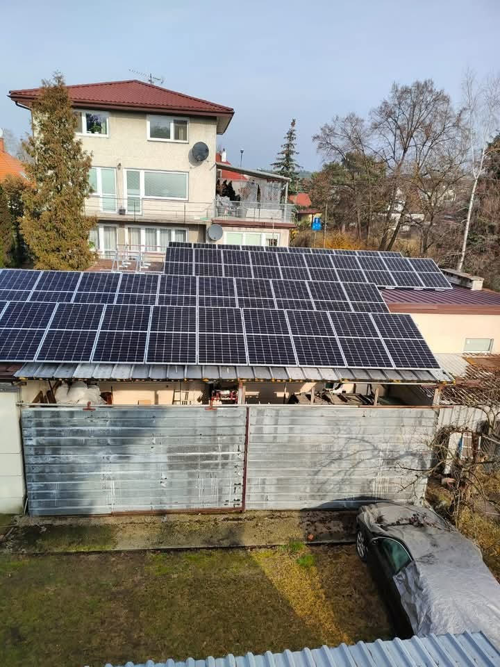 Instalacja paneli na dachu budynku gospodarczego, z widocznym samochodem przykrytym plandeką w ogrodzie i domem mieszkalnym w tle, uchwycone z góry w pochmurny dzień.