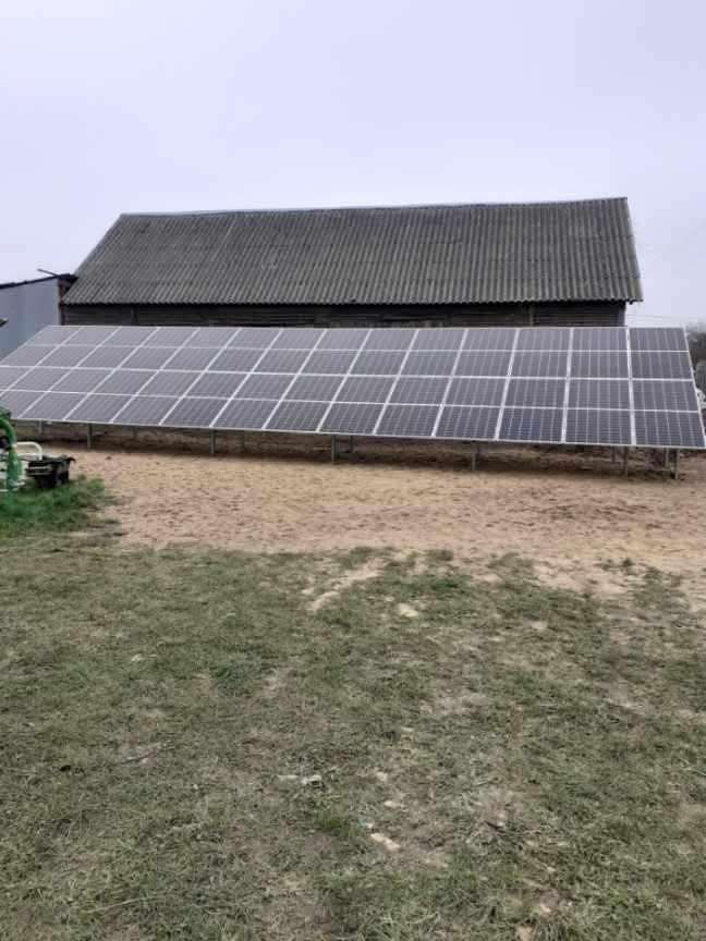 Rząd paneli słonecznych ustawionych na ziemi przed starym budynkiem gospodarczym z szarym dachem. Widoczny traktor z lewej strony. Pochmurne niebo.