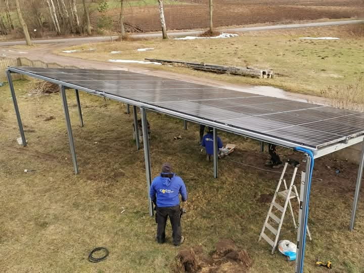 Montaż paneli słonecznych na metalowej konstrukcji w terenie niezabudowanym, widoczni dwaj instalatorzy w niebieskich bluzach z logo firmy, drabina oparta o konstrukcję, wiosenna sceneria.