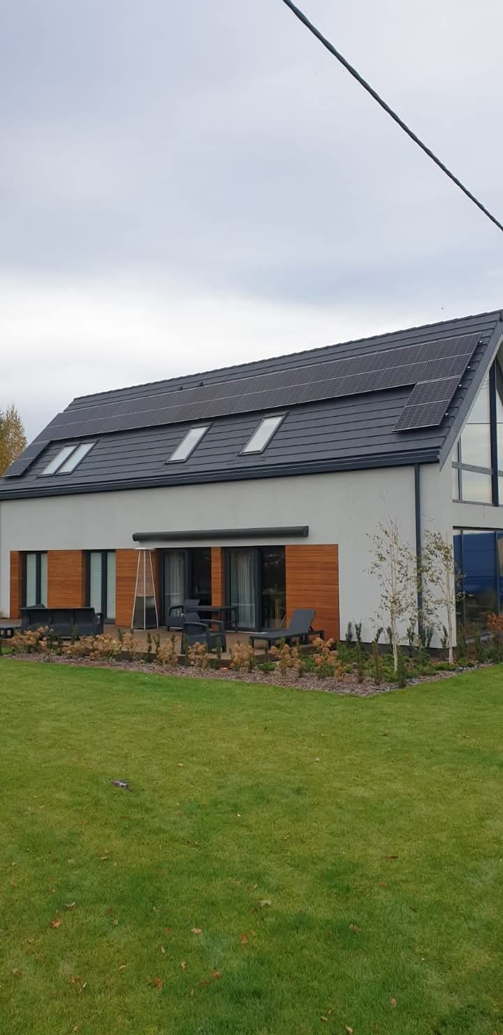 Nowoczesny dom jednorodzinny z panelami fotowoltaicznymi na dachu, widoczne okna dachowe i drewniana elewacja, otoczony zadbanym trawnikiem i roślinnością ozdobną.