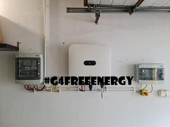 Biały inwerter centralny, dwie szare skrzynki bezpiecznikowe z widocznymi przewodami, umieszczone na białej ścianie garażu, z napisem #G4FREEENERGY.