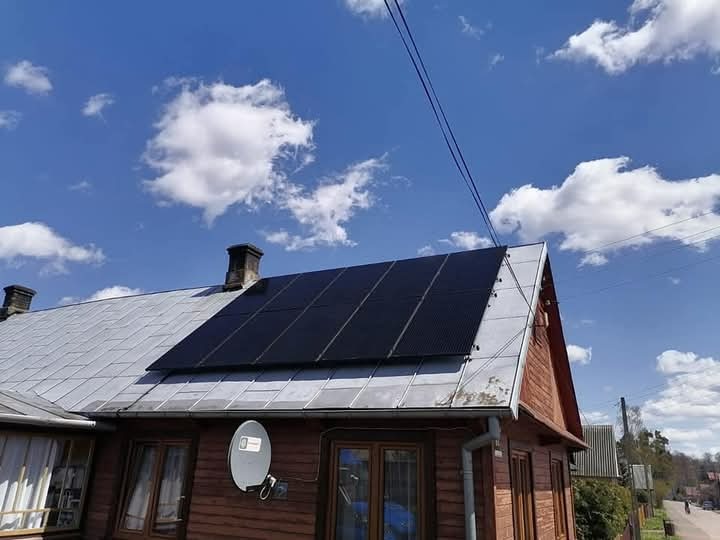 Panele słoneczne zamontowane na dachu drewnianego domu z widoczną anteną satelitarną i kominami, na tle błękitnego nieba z chmurami.
