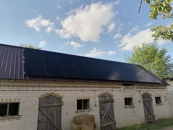 Częściowy widok instalacji paneli słonecznych na dachu ceglanego budynku gospodarczego z drewnianymi drzwiami, w tle błękitne niebo z chmurami i drzewa.