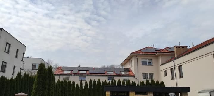 Domy jednorodzinne z panelami słonecznymi na dachach, widok z poziomu ulicy, pochmurne niebo w tle.