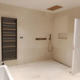 Jasna łazienka z beżowymi płytkami, czarnym grzejnikiem drabinkowym, minimalistyczną armaturą i otwartą kabiną prysznicową.