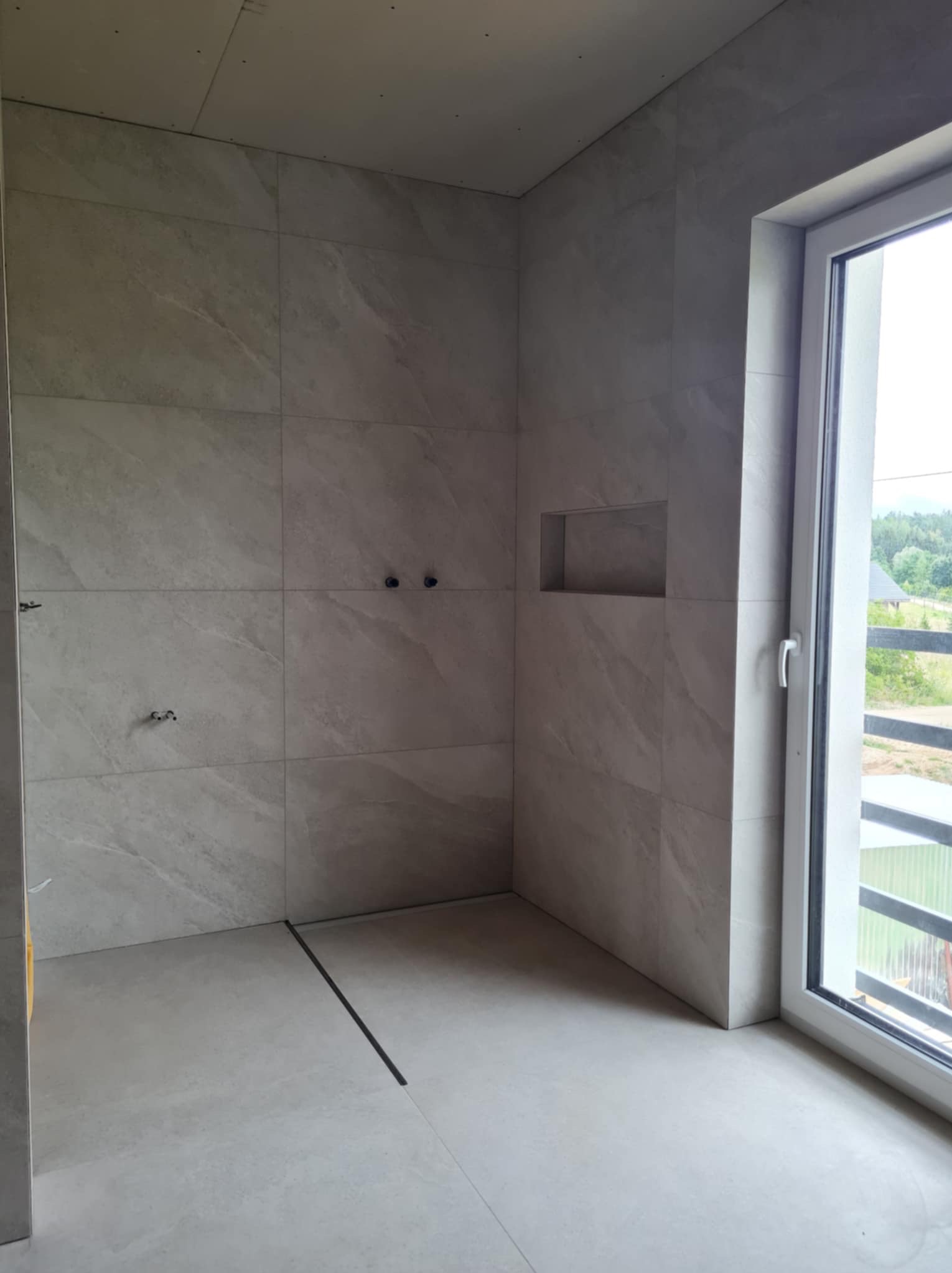 Wnętrze łazienki w trakcie wykańczania, widoczne szare płytki imitujące kamień na ścianach i podłodze, wnęka w ścianie, czarny odpływ liniowy w podłodze, okno z widokiem na zieleń, sufit z płyt...