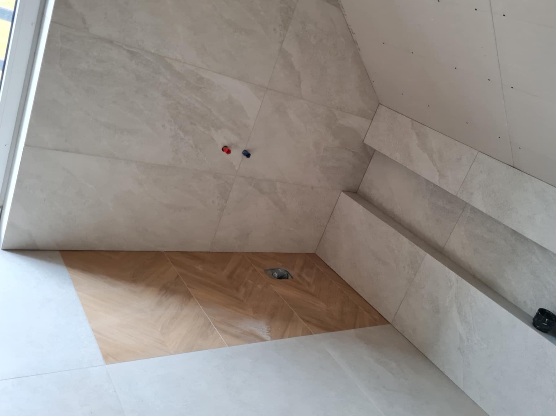 Fragment remontowanej łazienki z ułożonymi płytkami w odcieniach beżu i szarości, widoczne przyłącza wodne i odpływ pod prysznicem, ściany częściowo wykończone płytami kartonowo-gipsowymi.