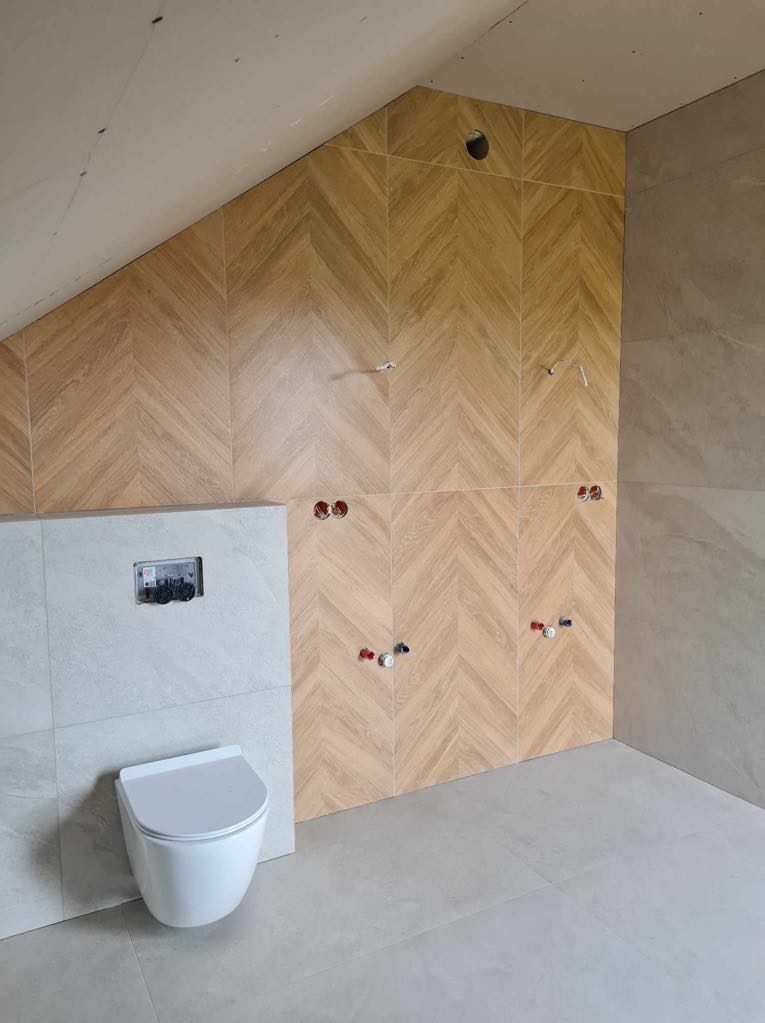 Montaż podtynkowej toalety wiszącej w łazience na poddaszu, ściany wyłożone płytkami imitującymi drewno ułożonymi w jodełkę, widoczne przyłącza wodne.