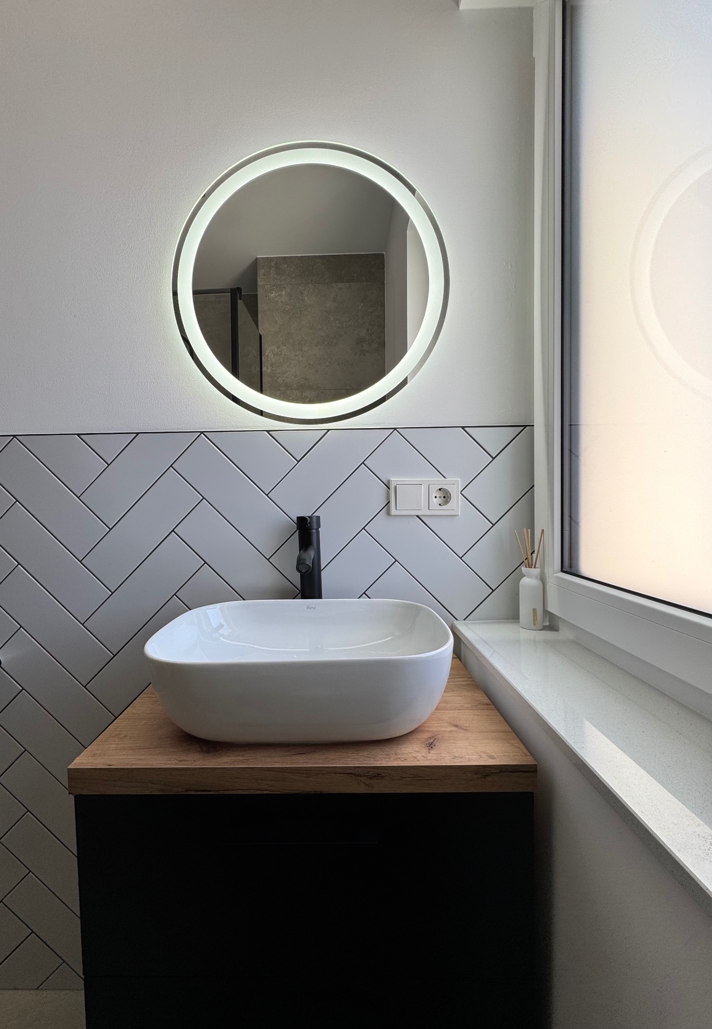 Nowoczesna łazienka z białą umywalką nablatową, czarną armaturą, okrągłym lustrem z podświetleniem LED i białymi płytkami ułożonymi w jodełkę.