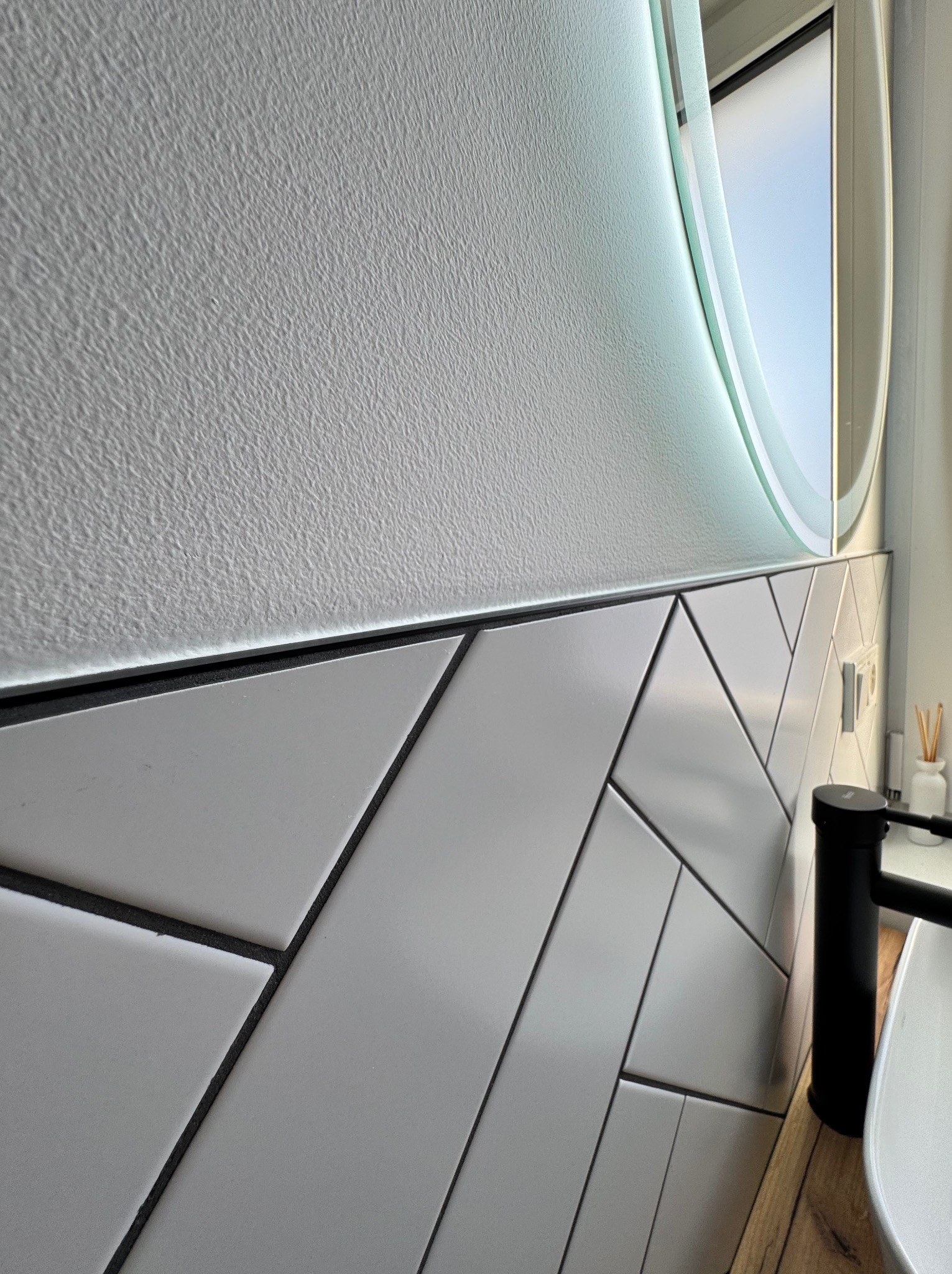 Ściana łazienki wykończona białymi, ukośnie ułożonymi płytkami z ciemną fugą, nad którymi znajduje się ściana z fakturą baranka i podświetlane okrągłe lustro.