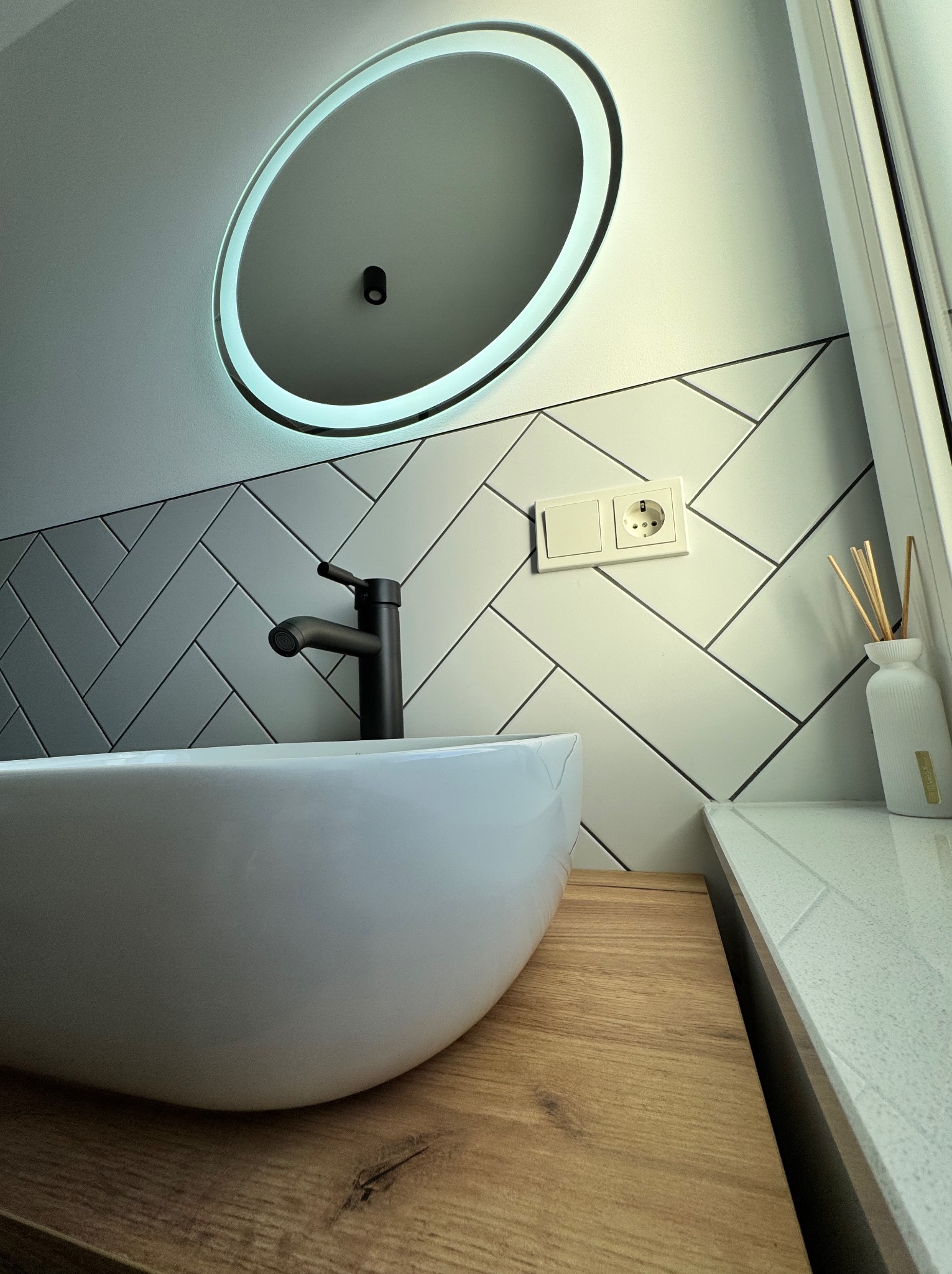Nowoczesna łazienka z owalną umywalką nablatową, czarną armaturą i podświetlanym lustrem, płytki ułożone w jodełkę, drewniany blat i biały dyfuzor zapachowy z patyczkami.