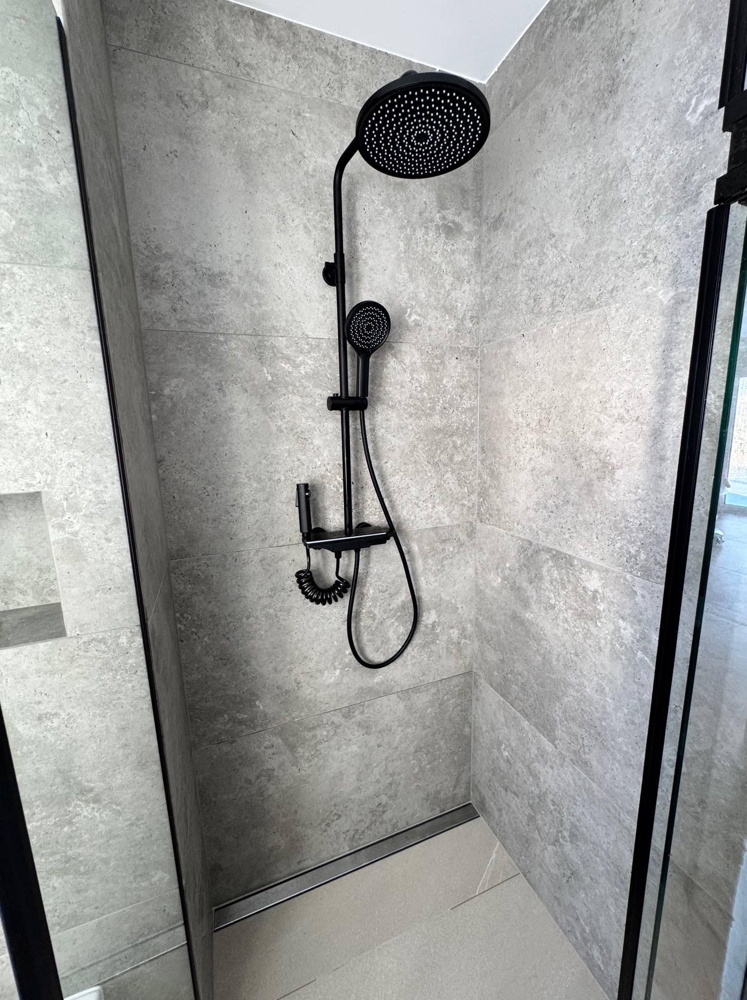 Nowoczesna kabina prysznicowa z szarymi płytkami imitującymi kamień, czarną armaturą i odpływem liniowym.