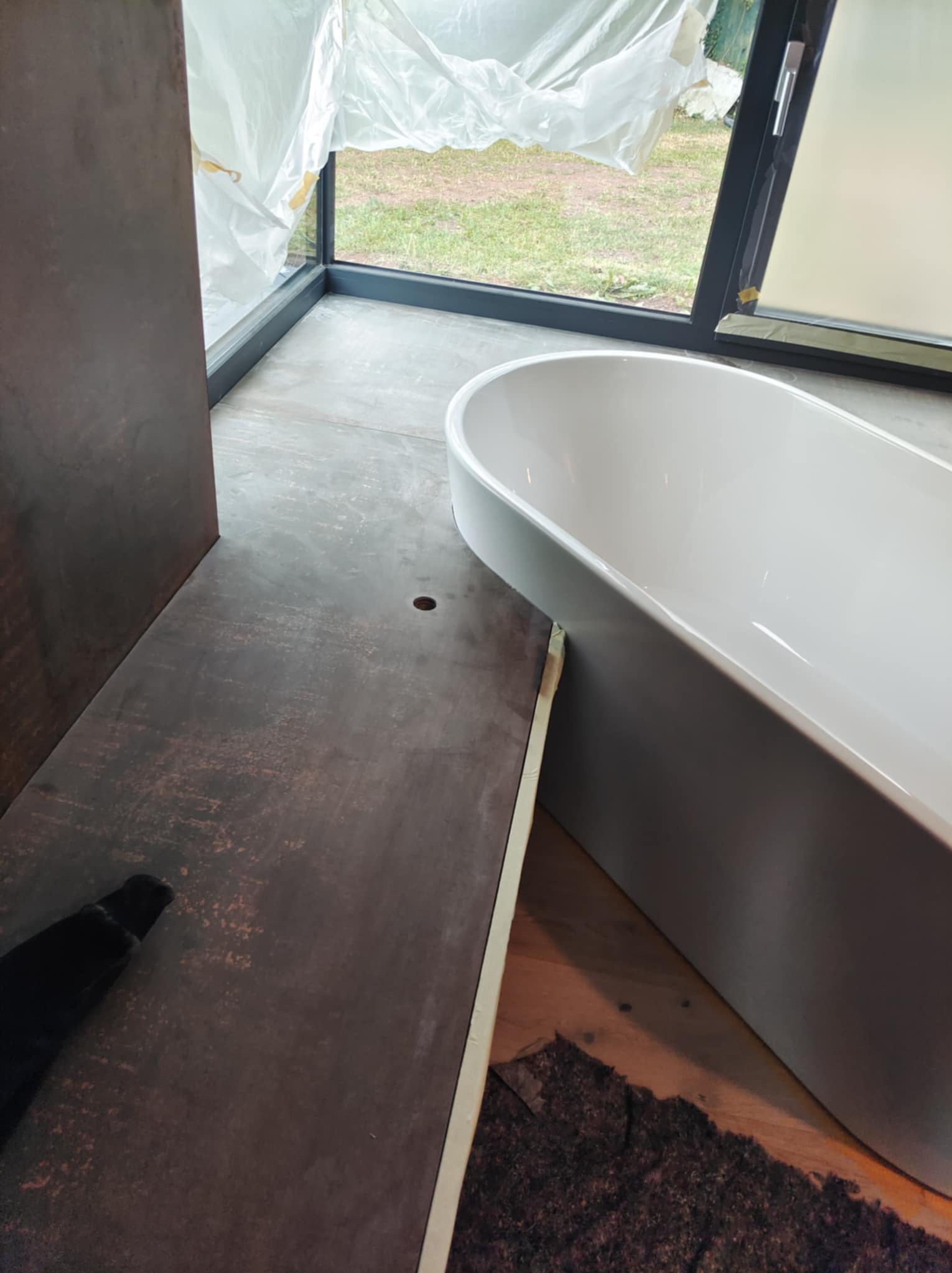 Wanna wolnostojąca w trakcie montażu w łazience z dużym oknem, zabezpieczonym folią. Widoczne przyłącze kanalizacyjne w podłodze, fragment instalacji oraz zabezpieczenia z kartonu i taśmy malarskiej.