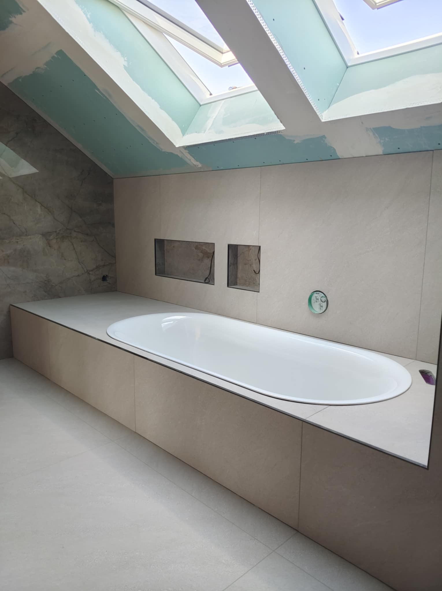 Łazienka w trakcie wykańczania: biała wanna obudowana płytkami, dwa wnęki w ścianie, płyty kartonowo-gipsowe na skosach sufitu z oknami dachowymi.