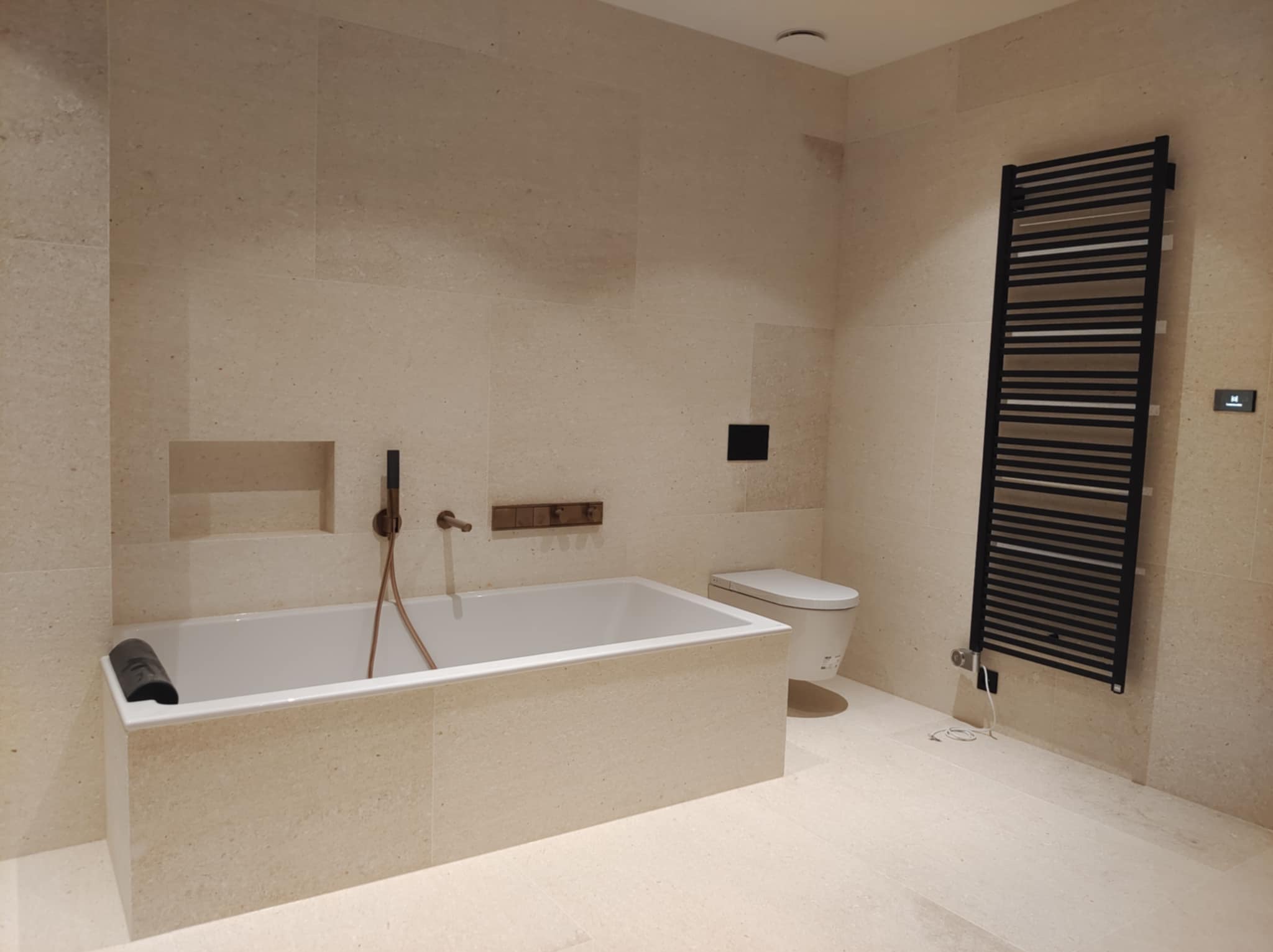 Jasna łazienka z wanną obłożoną płytkami imitującymi kamień, czarnym grzejnikiem drabinkowym i toaletą w tle.