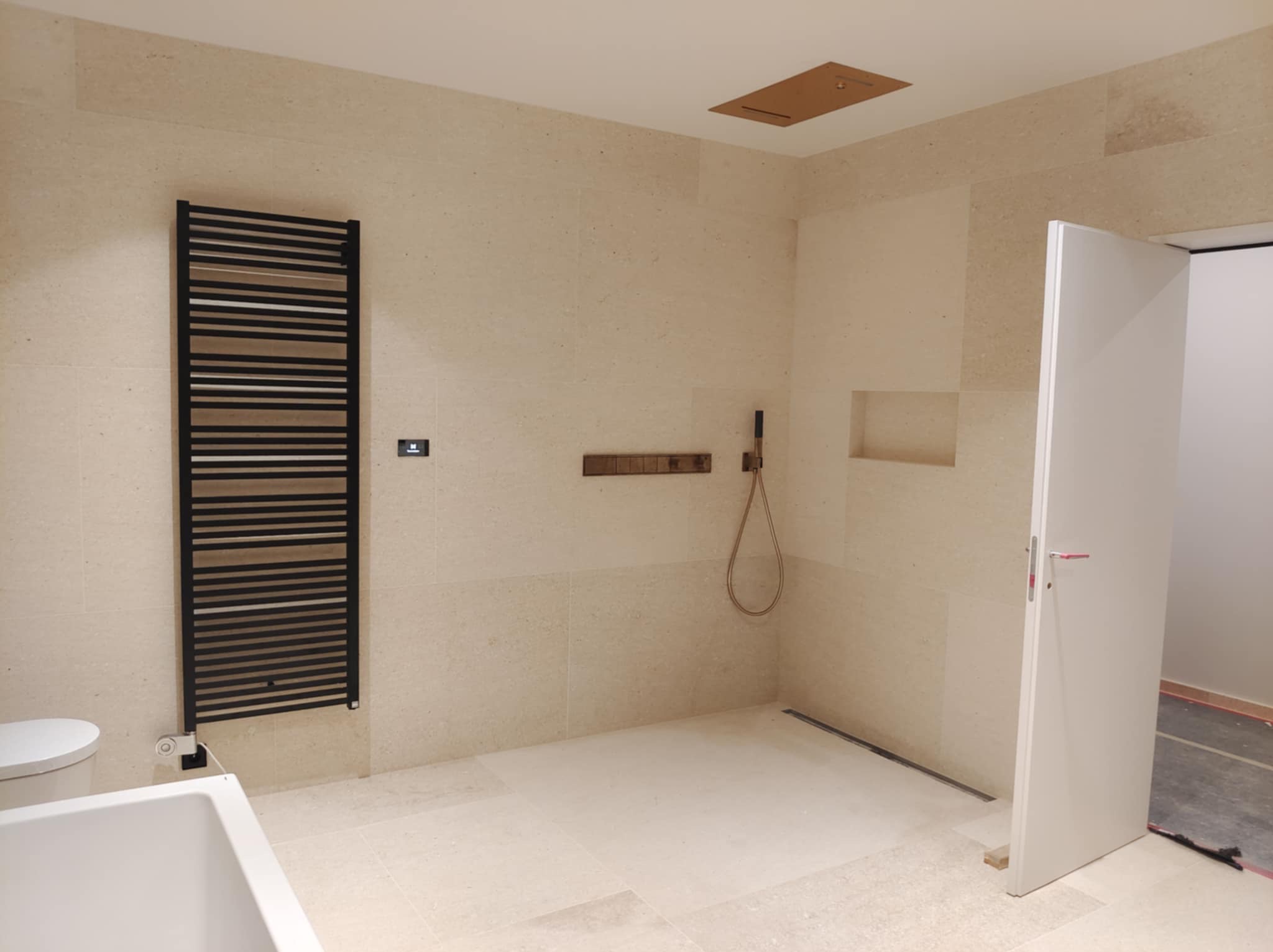 Jasna łazienka z beżowymi płytkami, czarnym grzejnikiem drabinkowym, minimalistyczną armaturą i otwartą kabiną prysznicową.