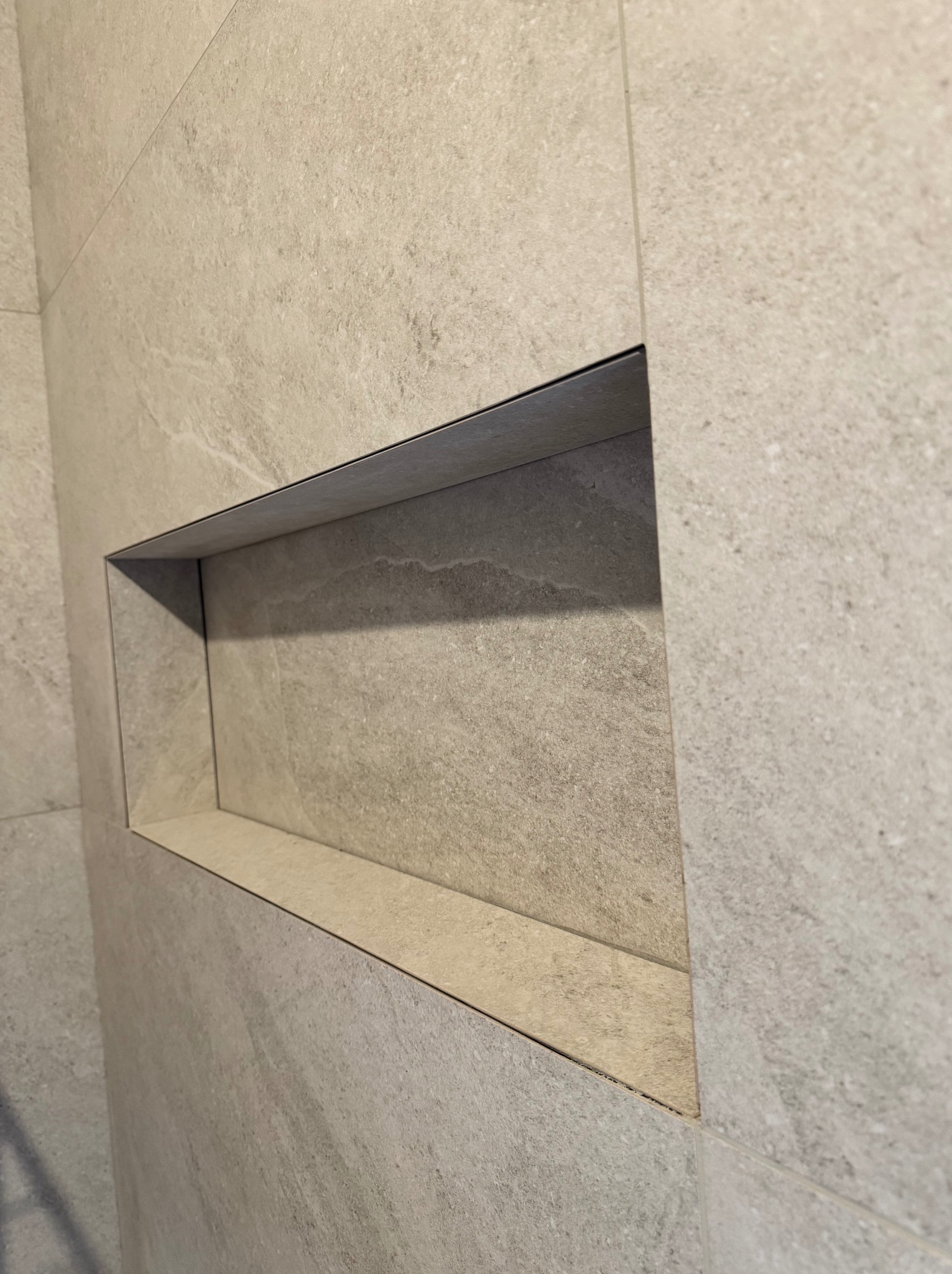 Wnęka w ścianie wyłożonej płytkami ceramicznymi w odcieniach beżu i szarości, wykończona ciemnoszarą listwą, oświetlona naturalnym światłem padającym z góry.